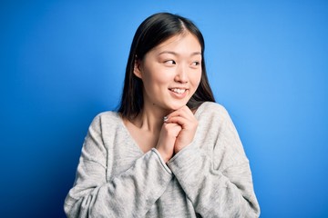 Young beautiful asian woman wearing casual sweater standing over blue isolated background laughing nervous and excited with hands on chin looking to the side