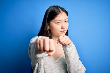 Fototapeta premium Young beautiful asian woman wearing casual sweater standing over blue isolated background Punching fist to fight, aggressive and angry attack, threat and violence