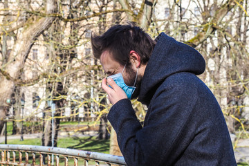 Fototapeta na wymiar Handsome young European man coughing on the street with a medical face mask on. Closeup of a 35-year-old male in a respirator to protect against infection with influenza or coronavirus (Covid-19)