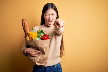 Young asian woman holding paper bag of fresh healthy groceries over yellow isolated background pointing displeased and frustrated to the camera, angry and furious with you