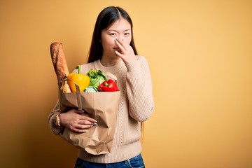Young asian woman holding paper bag of fresh healthy groceries over yellow isolated background smelling something stinky and disgusting, intolerable smell, holding breath with fingers on nose