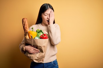 Young asian woman holding paper bag of fresh healthy groceries over yellow isolated background Yawning tired covering half face, eye and mouth with hand. Face hurts in pain.