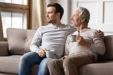 Obraz na płótnie Canvas Thoughtful elderly father and adult son sit relax on couch in living room drink tea look in distance dreaming, pensive dreamy mature adult and grown-up child rest on sofa at home visualizing together