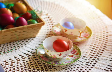 Easter colorful eggs, flowers and greens, white tea-set with eggs inside the cups with butterflies thin porcelain, on a white rounded knitted tablecloth. Country cozy style - happy holiday!