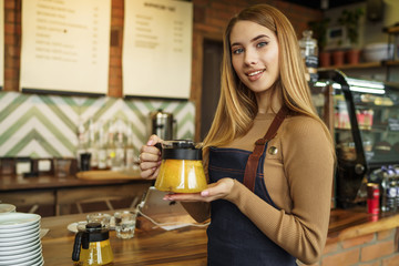 Cute young beautiful girl, caucasian barista in apron smiling at camera in coffee shop counter with mandarin tea in kettle