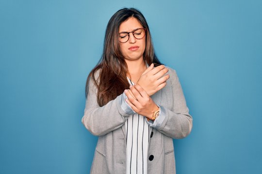 Young hispanic business woman wearing glasses standing over blue isolated background Suffering pain on hands and fingers, arthritis inflammation