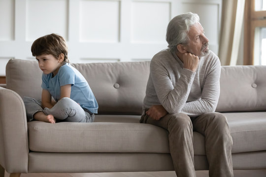 Upset offended little boy and senior grandfather sit separate on couch at home avoid talking after fight, sad stubborn mature grandparent and small grandson ignore each other, generation gap concept