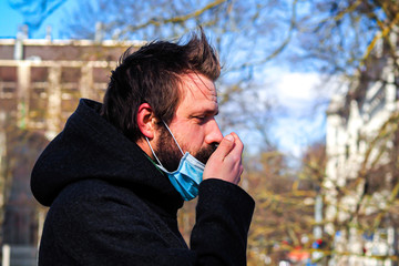 Handsome young European man coughing on the street with a medical face mask on. Closeup of a 35-year-old male in a respirator to protect against infection with influenza or coronavirus (Covid-19)