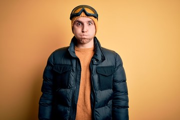 Young handsome caucasian man wearing hat, coat and ski glasses for winter and snow weather puffing cheeks with funny face. Mouth inflated with air, crazy expression.