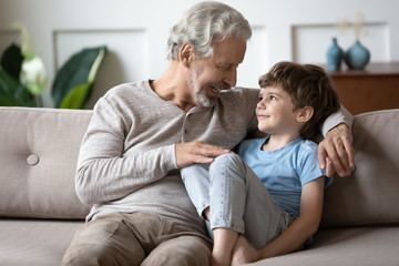 Happy loving mature grandfather sit rest on sofa in living room enjoy weekend with little grandchild, smiling senior grandparent relax on couch at home talk chat with cute small preschooler grandson