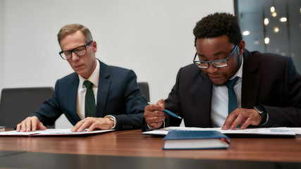 Paying attention to every detail. Two focused african and caucasian businessman in formal wear analyzing some documents while sitting at the office table in the modern office