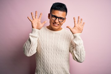 Young handsome latin man wearing white casual sweater and glasses over pink background showing and pointing up with fingers number ten while smiling confident and happy.