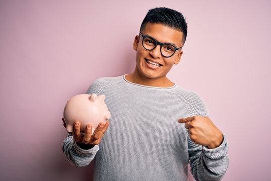 Young handsome latin man holding piggy bank over isolated pink background with surprise face pointing finger to himself