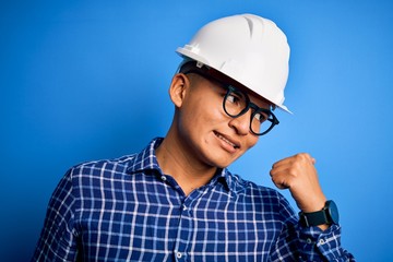 Young handsome engineer latin man wearing safety helmet over isolated blue background smiling with happy face looking and pointing to the side with thumb up.