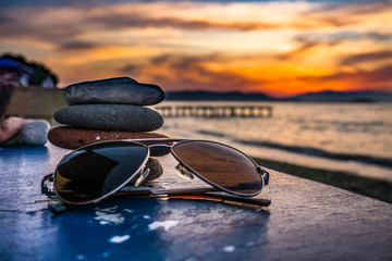 Fototapeta na wymiar Sunglasses closeup on the table on beach towards sunset over sea and coast pier. Holiday, vacation and relaxation conceptual photography. Retro or modern sunglasses at seaside or shore.