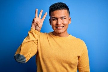 Young handsome latin man wearing yellow casual sweater over isolated blue background showing and pointing up with fingers number three while smiling confident and happy.