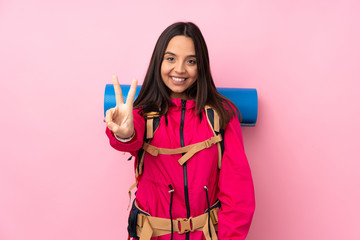 Young mountaineer girl with a big backpack over isolated pink background smiling and showing victory sign