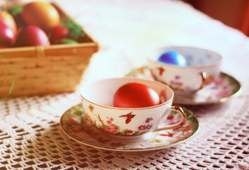 Easter colorful eggs, flowers and greens, white tea-set with eggs inside the cups with butterflies thin porcelain, on a white rounded knitted tablecloth. Country cozy style - happy holiday!