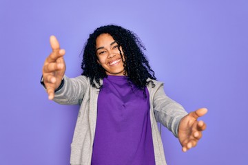 Young african american sporty woman wearing casual sweatshirt over purple background looking at the camera smiling with open arms for hug. Cheerful expression embracing happiness.