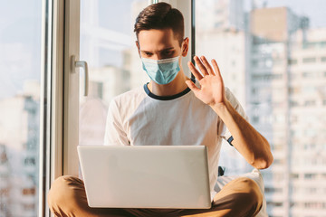 Student freelancer having online business meeting and sitting on windowsill. Young businessman in medical protective face mask chatting on laptop while working on computer at home. Online conference