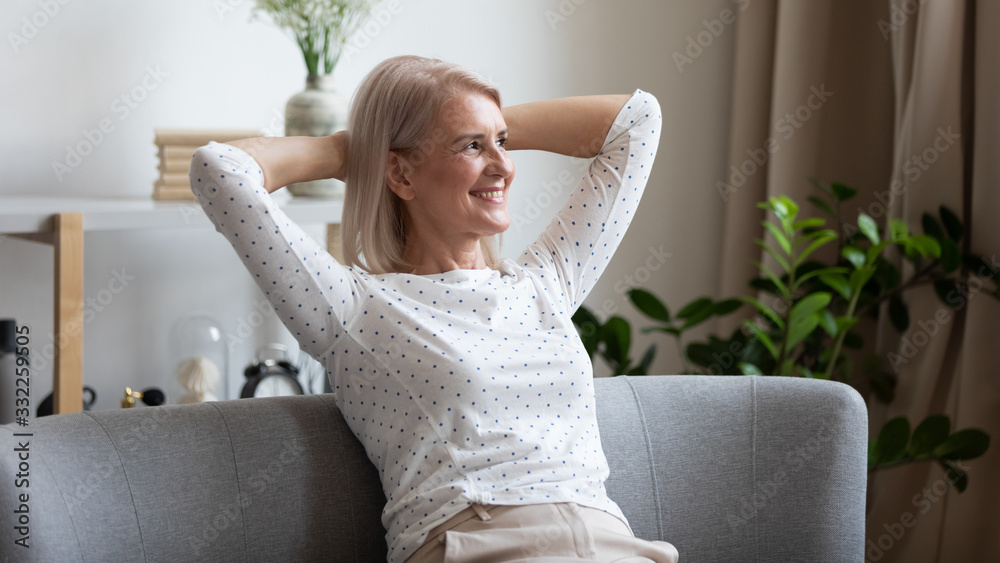 Wall mural pleasant smiling middle aged woman relaxing on cozy coach in modern living room, looking away. happy