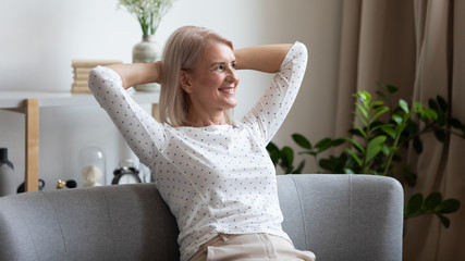 Pleasant smiling middle aged woman relaxing on cozy coach in modern living room, looking away....