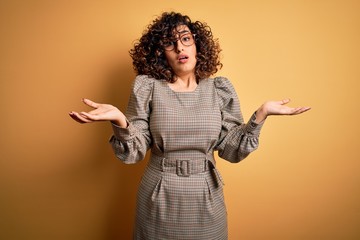 Beautiful arab business woman wearing dress and glasses standing over yellow background clueless...