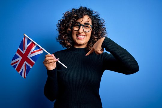 Young beautiful curly arab woman wearing glasses holding uk flag over blue background with surprise face pointing finger to himself - Powered by Adobe