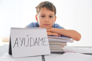 Tired frustrated boy sitting at the table with many books, exercises books. Spanish word Auydame -...