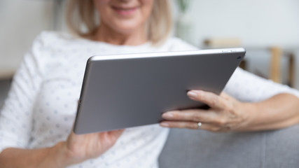 Close up pleasant elder female retiree holding digital tablet, sitting on couch at home. Positive middle aged woman searching information or shopping online. Older generation easy technology usage.