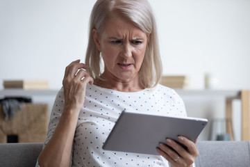 Frowning middle aged woman looking at computer tablet, irritated by spam email or bad gadget work,...