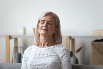 Peaceful middle aged blonde woman leaning on sofa, relaxing with closed eyes. Calm mature retired lady resting in silence, meditating, visualizing future, enjoying weekend leisure time alone at home.