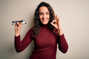 Young beautiful woman with curly hair holding glasses over isolated white background doing ok sign with fingers, excellent symbol