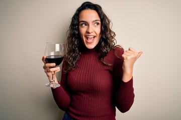 Young beautiful woman with curly hair drinking glass of red wine over white background pointing and...