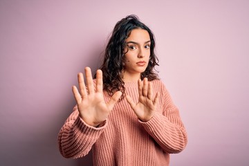 Young beautiful woman with curly hair wearing casual sweater over isolated pink background Moving away hands palms showing refusal and denial with afraid and disgusting expression. Stop and forbidden.