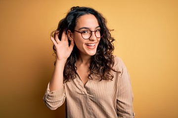 Beautiful woman with curly hair wearing striped shirt and glasses over yellow background smiling with hand over ear listening an hearing to rumor or gossip. Deafness concept.