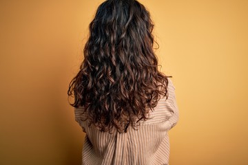 Beautiful woman with curly hair wearing striped shirt and glasses over yellow background standing backwards looking away with crossed arms