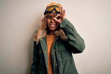 Young african american skier woman with curly hair wearing snow sportswear and ski goggles doing ok gesture with hand smiling, eye looking through fingers with happy face.