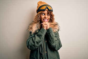 Young african american skier woman with curly hair wearing snow sportswear and ski goggles laughing nervous and excited with hands on chin looking to the side