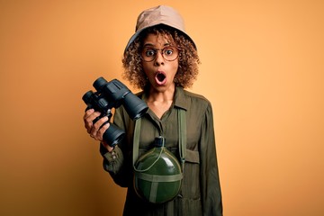 African american tourist woman wearing explorer hat and water canteen holding binoculars scared in...
