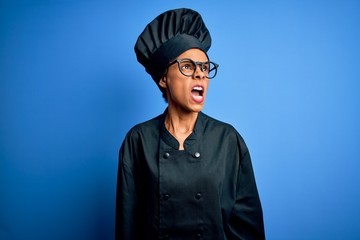 Young african american chef woman wearing cooker uniform and hat over blue background angry and mad screaming frustrated and furious, shouting with anger. Rage and aggressive concept.