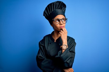 Young african american chef woman wearing cooker uniform and hat over blue background with hand on chin thinking about question, pensive expression. Smiling with thoughtful face. Doubt concept.