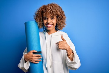 Young african american woman doing sport holding yoga mat over isolated blue background with surprise face pointing finger to himself