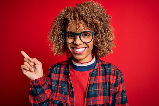 Young beautiful african american woman wearing casual shirt and glasses over red background with a big smile on face, pointing with hand and finger to the side looking at the camera.