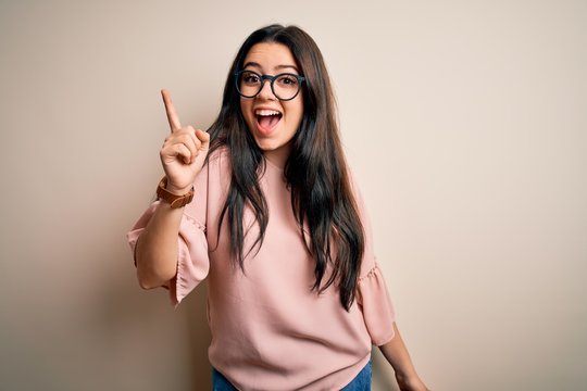 Young brunette elegant woman wearing glasses over isolated background pointing finger up with successful idea. Exited and happy. Number one.