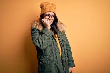 Young brunette woman wearing glasses and winter coat with hat over yellow isolated background looking stressed and nervous with hands on mouth biting nails. Anxiety problem.