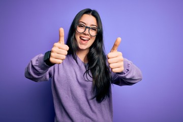 Young brunette woman wearing glasses over purple isolated background approving doing positive gesture with hand, thumbs up smiling and happy for success. Winner gesture.