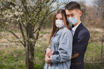 Asian guy with a European girl in medical masks on the background of flowering trees hug and kiss