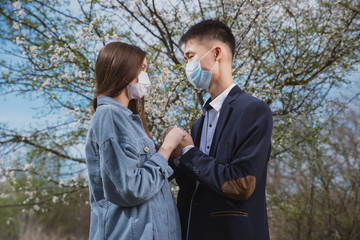 Asian guy with a European girl in medical masks on the background of flowering trees hug and kiss