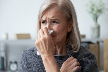 Head shot close up unhealthy older woman covered in plaid, using paper tissue, wiping runny nose. Unhappy mature lady caught cold or suffering from seasonal allergy. Healthcare vaccination concept.